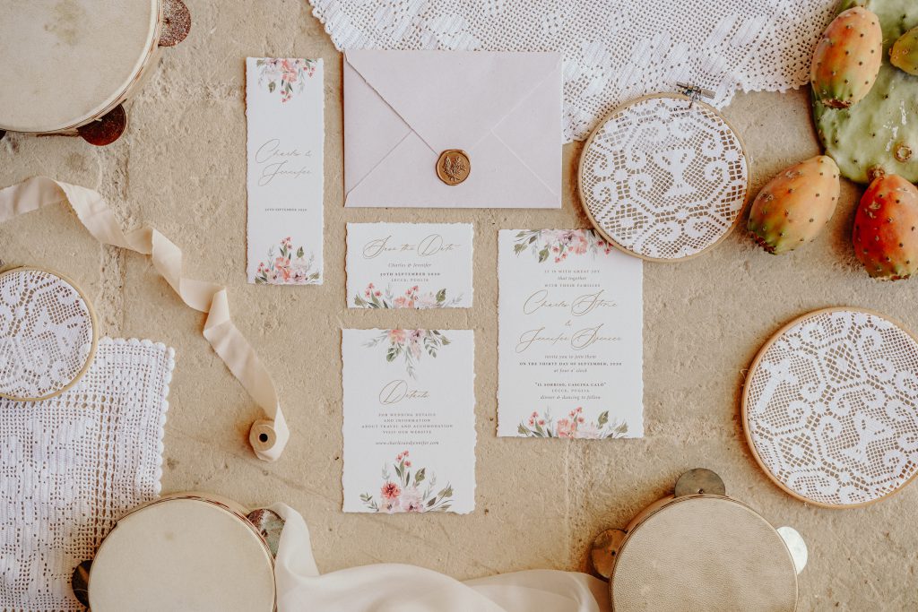 Partecipazioni e save the date con fiori acquerellati, ceralacca dorata, tamburelli e pizzo su una pietra chiara