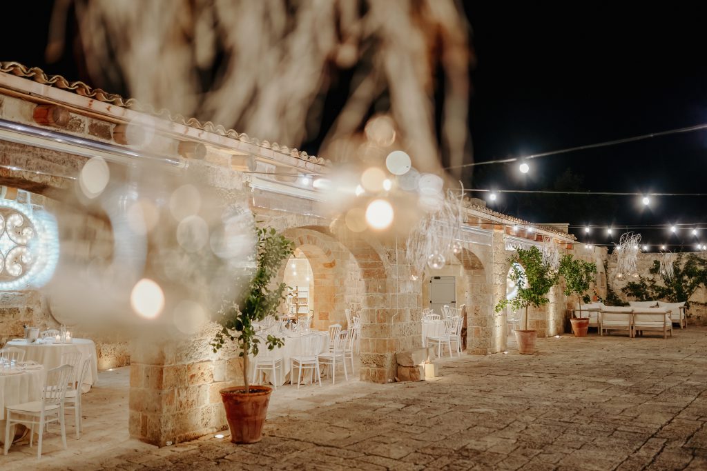 Sposi in una sala a volta in pietra, accanto a un tavolo imperiale bianco decorato di fiori e lampadine
