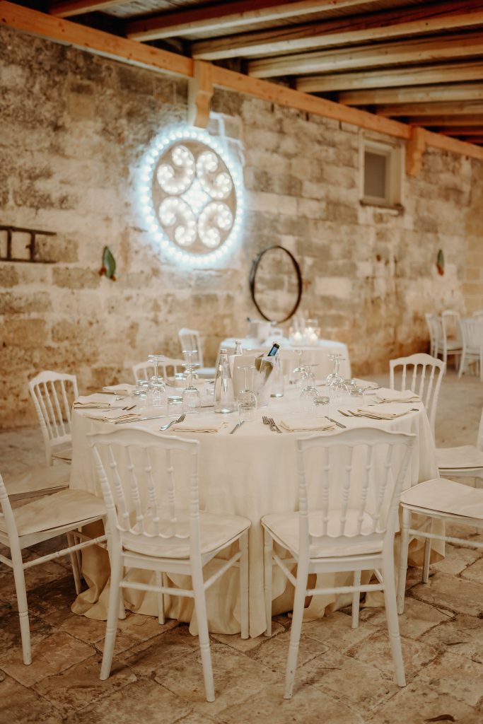 Sala con muri in pietra a vista e travi di legno, tavolo rotondo apparecchiato e rosone illuminato alla parete