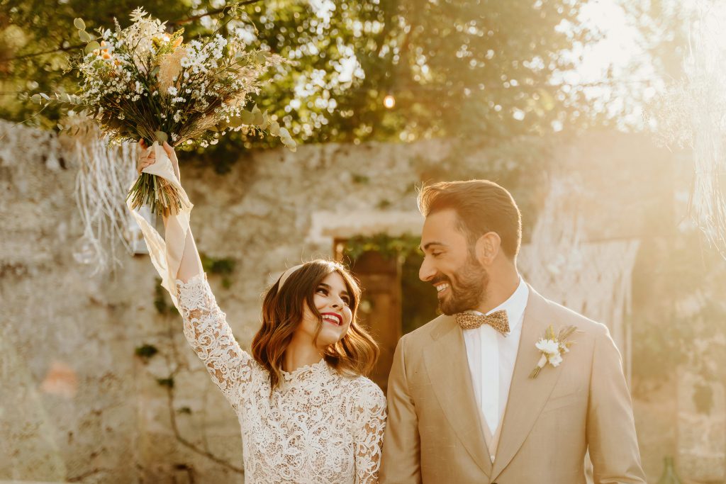 Sposi che ridono davanti a un muro in pietra al tramonto, lei alza il bouquet di fiori secchi