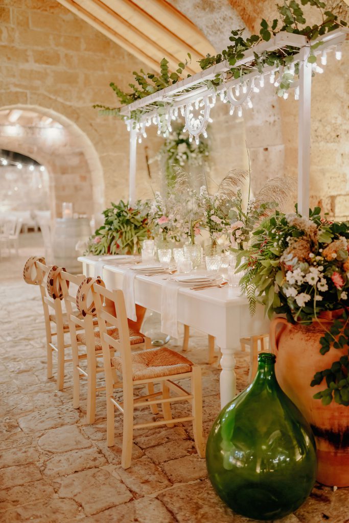 Tavolo imperiale apparecchiato di bianco sotto la volta in pietra, con fiori di campo e luci appese